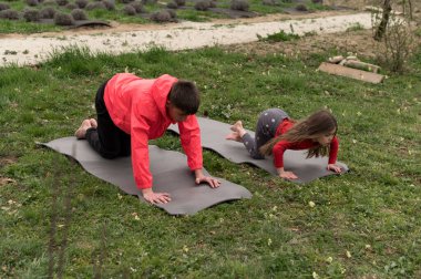 Huzurlu bir bahar gününün tadını çıkaran bir baba ve kızı yemyeşil bir bahçede minderlerde yoga yapıyor..