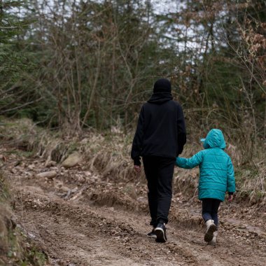 Parlak bir turkuaz ceket giyen bir çocuk, dağılmış taşlar ve çalılarla kaplı engebeli bir orman yolunda yürürken siyah giyinmiş bir yetişkinin elini sıkıca tutuyor..