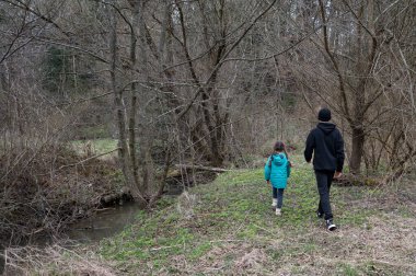 Baharın başlarında, yeşil ceketli bir çocuk, tomurcuklanan ağaçlar ve yakındaki bir derenin yumuşak sesleri ile çevrili, dingin bir orman yolunda bir yetişkinin yanında yürür..