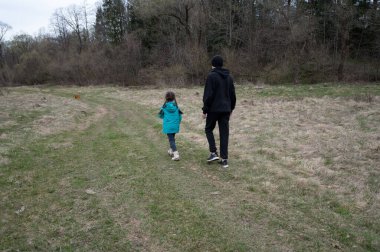 Parlak bir turkuaz ceket giyen bir çocuk bulutlu bir günde, yemyeşil ve çıplak ağaçların arasında dolambaçlı bir patikada bir yetişkinin yanında neşeyle yürüyor..