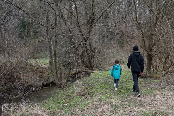Baharın başlarında, yeşil ceketli bir çocuk, tomurcuklanan ağaçlar ve yakındaki bir derenin yumuşak sesleri ile çevrili, dingin bir orman yolunda bir yetişkinin yanında yürür..