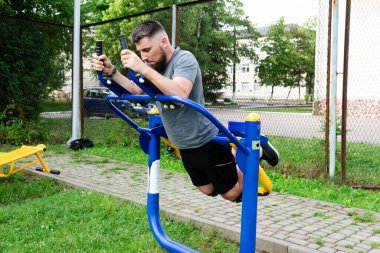 Sakallı bir adam dışarıda spor aletleri üzerinde sırt egzersizi yapıyor. Odaklanmış ve kararlı, park ortamında sağlıklı bir egzersizin tadını çıkarıyor..