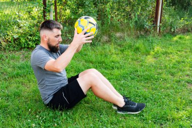 Sakallı bir adam çimenli bir alanda oturur, sarı ve siyah bir futbol topunu dikkatle inceler. Spor kıyafetleri giymiş, spor aktivitelerine ara verdiğini söylüyor..