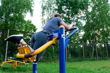 Bir kadın sağlıklı ve aktif bir yaşam tarzı sergilemek için açık hava fitness makinesi kullanıyor. Arka planda yeşil çimenler ve ağaçlar olan bir park..