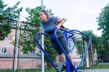 Bir kadın dışarıda spor aletleri kullanarak üst vücut egzersizi yapıyor. Atletik kıyafetler giyiyor ve egzersizine odaklanmış görünüyor. Ortam halka açık bir park ya da açık bir alan..