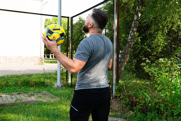 Gri tişörtlü ve siyah şortlu sakallı bir adam sarı bir futbol topuyla hokkabazlık yapıyor. Dışarıda, tel örgülerin yanında güneşli bir günün tadını çıkarıyor..