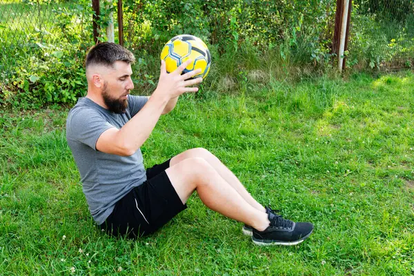 Sakallı bir adam çimenli bir alanda oturur, sarı ve siyah bir futbol topunu dikkatle inceler. Spor kıyafetleri giymiş, spor aktivitelerine ara verdiğini söylüyor..