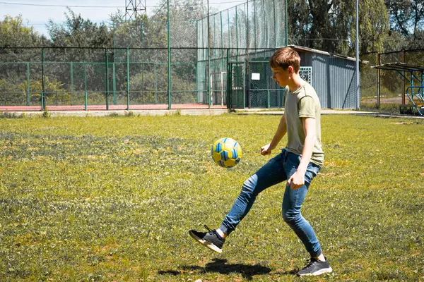 Genç bir insan çimenli bir alanda bir futbol topuyla ustaca hokkabazlık yapıyor. Arka planda çitli bir spor salonu görülebilir..