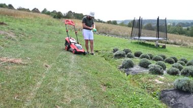 Bir adam çim biçme makinesini çimlerin kesildiği yere götürüyor, çimler için bahçe aletleri..
