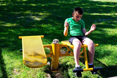 Bir çocuk sarı spor aletlerine oturup mekik çekiyor. Yeşil bir gömlek ve şort giyiyor. Sahne parlak ve güneşli, yemyeşil otlarla dolu..