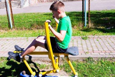 Genç bir çocuk açık hava egzersiz ekipmanları kullanarak bacak presine odaklanırken görülüyor. Spor giysileri giyiyor, güneşli bir fiziksel aktivite gününün tadını çıkarıyor..