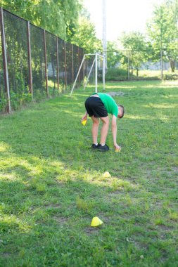 Yeşil gömlekli bir çocuk çimenli bir spor sahasına sarı koni koymak için eğilir. Arka planda bir futbol golü görünür. Atletik antrenman için hazırlanıyor..