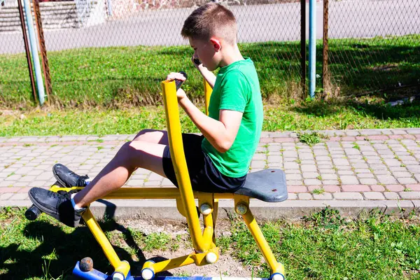 Genç bir çocuk açık hava egzersiz ekipmanları kullanarak bacak presine odaklanırken görülüyor. Spor giysileri giyiyor, güneşli bir fiziksel aktivite gününün tadını çıkarıyor..