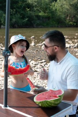 Mutlu bir kız ve babası nehir kenarındaki piknik sırasında karpuz dilimlerini paylaşırlar. Onlar bir plaj şemsiyesinin altında bir masada oturuyorlar, yaz gününün tadını çıkarıyorlar..