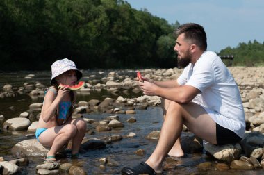 Bir adam ve genç bir kız sığ bir nehrin kenarındaki kayalarda oturmuş, mutlu bir şekilde karpuz dilimleri yiyorlar. Sahne huzurlu ve huzurlu, güneş ışığıyla yıkanmış..