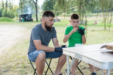 Bir adam ve çocuk portatif bir parkta oturuyorlar, siyah eldiven giyerek yemek hazırlıyorlar. Rahatlamış görünüyorlar ve piknik tarzında yemek yiyorlar..
