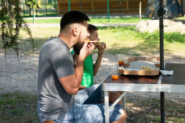 Bir adam ve bir çocuk parkta küçük bir katlama masasında oturup pizza dilimleri yiyip meyve suyu içiyorlar. Sahne rahatlatıcı ve neşeli. Yürek ısıtan bir baba-oğul anı..