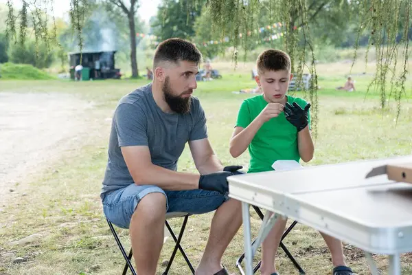 Bir adam ve çocuk portatif bir parkta oturuyorlar, siyah eldiven giyerek yemek hazırlıyorlar. Rahatlamış görünüyorlar ve piknik tarzında yemek yiyorlar..