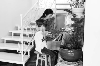 Bir çocuğun klavye çalmasını öğretirken çekilmiş siyah beyaz bir fotoğraf. Evcil bitkilerle çevrili bir merdivenin yanındaki küçük bir tabureye yerleştirilmişler..