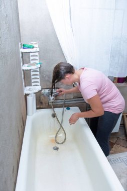 Bu fotoğraf, saçlarını eski bir küvette, duş hortumu ve metal duş başlığıyla yıkayan bir kadını gösteriyor. Banyo eskimiş bir küvet, beton duvarlar ve açık bir pencereyle basit görünüyor..