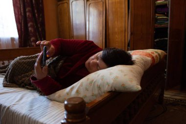 Rahat bir kadın, cep telefonunu kullanarak yatakta dinleniyor. Dekorasyonda rahat ahşap mobilyalar ve rahatlatıcı bir battaniye yer alıyor ve sıcak bir ortamda dinlenme ve bağlantı sahnesi yaratıyor..