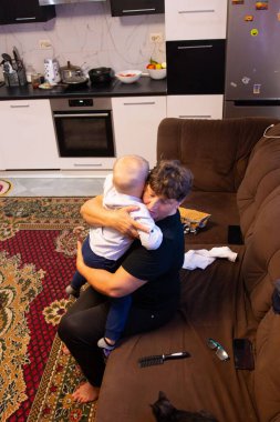 Yürek ısıtan bir görüntü, kucağında bir çocuk tutan bir yetişkini yakalar. Dekor, arka planda bir mutfak görüntüsü olan rahat bir yaşam alanı. Bu da bir aile evi olduğunu gösteriyor..