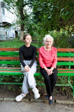 Bereketli bir şehir bahçesinde, genç bir kadın ve yaşlı bir bayan renkli bir park bankında birbirlerine eşlik ederek, sakin bir öğleden sonra gökyüzünün altında sıcak bir gülümseme takınırlar..
