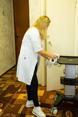 Sağlık görevlisinin tıbbi bir cihazı temizlemek için kağıt havlu kullandığını gösteren bir görüntü. Beyaz önlük, siyah pantolon ve spor ayakkabı giyiyor. Steril bir ortam sağlıyor..