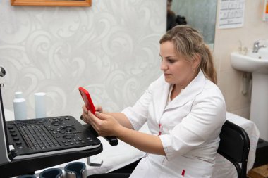 Bir tıp uzmanı, sonografi makinesinin yanındaki cep telefonunda bilgileri kontrol ediyor. Dekorasyon büyük olasılıkla bir doktor muayenehanesi ya da klinik, ve yanında temel tıbbi tesisler bulunuyor..