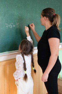 Bir öğretmen, sınıfta yeşil bir karatahta üzerinde matematik problemi çözen bir öğrenciye rehberlik eder. Eğitim ve matematik beceri geliştirme üzerine odaklanarak, kurum eğitici nitelikte..