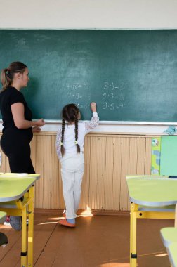 Bir öğretmen sınıftaki yeşil bir karatahtaya matematik problemleri yazan genç bir öğrenciye talimat verir. Sahne ön planda okul sıralarıyla aydınlık, öğrenme ve talimatları vurguluyor.