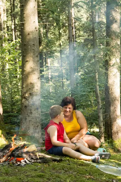 Bir kadın ve küçük bir çocuk, güneşli bir ormanda kamp ateşinin yanında oturuyorlar. Sohbete dalıyorlar, huzurlu ormanlık alanların tadını çıkarıyorlar..