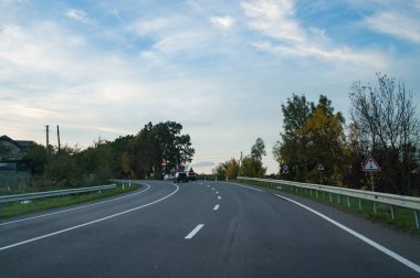 Sonbahar renklerinde ağaçlarla çevrili kıvrımlı bir yol huzurlu bir kırsal alana götürüyor, açık mavi gökyüzü altında keşif yapmaya davet ediyor..