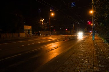Uzaktaki farlar karanlığı delip geçerken yağmur damlaları kaldırımda parlıyor. Sessiz cadde, sokak lambalarının yumuşak parıltısıyla canlıdır. Gece gezginlerini davet eder..