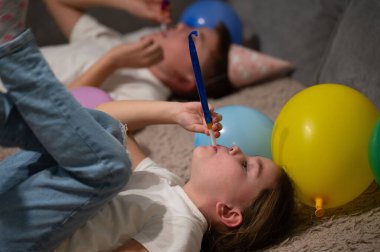 İki neşeli çocuk yumuşak bir yüzeye uzanmış, parti şapkaları takmış ve neşeli boynuzlarını üflüyorlar, etrafları canlı balonlarla çevrili, evde bir kutlamanın keyfini yakalıyorlar..