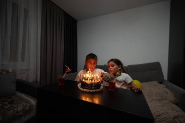İki çocuk, sevgi dolu, yumuşak ışıklı bir oturma odasında mumlarla süslenmiş güzel bir pastaya yaslanmış, samimi bir doğum günü anını paylaşıyor..