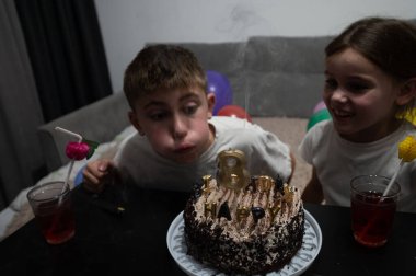 Balonlar ve içkiler şenlikli bir ortam hazırlarken iki çocuk güzel dekore edilmiş çikolatalı pastanın etrafında toplanıp mumları söndürürken kutlama başlıyor..