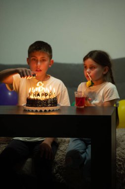 İki çocuk, güzel dekore edilmiş bir pastanın etrafında yanan mumlarla sıcak ve davetkar bir atmosferde doğum günü kutlamasının tadını çıkarıyorlar..