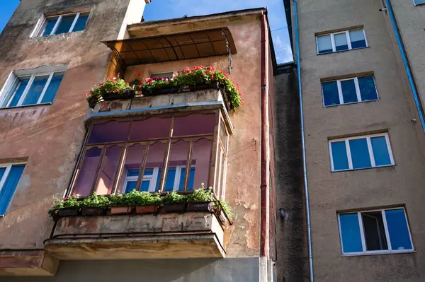 Yıpranmış bir bina, parlak mavi gökyüzünün altındaki komşu yapılarla çelişen renkli çiçeklerle dolu sevimli bir balkon sergiliyor..