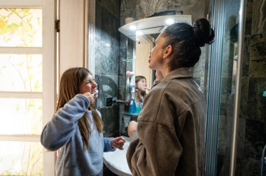 Genç bir kız mutlu bir şekilde dişlerini ablasıyla birlikte hoş bir banyoda fırçalıyor. Yumuşak ışık onların yakın ilişkilerini yansıtan sıcak bir atmosfer yaratıyor..