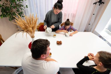 Güneşli bir odada, bir aile birlikte çay içer. Yetişkinler gülümsemeyi paylaşırken, bir çocuk merakla izliyor, bağlantı ve neşe dolu sıcak bir atmosfer yaratıyor..