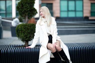 Şık bir beyaz önlük giymiş kendine güvenen bir kadın güneşli bir günde, yemyeşil ve şık kentsel tasarımlarla çevrili çağdaş bir bankta zarafetle oturur..