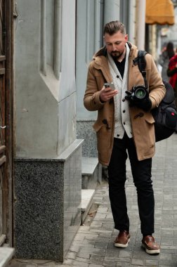 Sıcak bir palto giymiş bir adam kamerasını tutarken akıllı telefonunu kontrol eder. Kış öğleden sonraları canlı şehir yaşamını dikkatle izler ve yaratıcı enerji yayar..