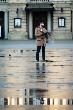 Sakin bir şehir meydanında, bir adam telefonuna dalmış bir şekilde dikilirken yağmur suları parıldayan yansımalar yaratıyor, etrafında güvercinler huzur içinde dolanıyor..