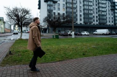 Islak kaldırımda yürürken, kahverengi paltolu bir adam gri gökyüzünün altında canlı bir şehirde çağdaş binaların önünden geçerken bir evrak çantası taşıyor..