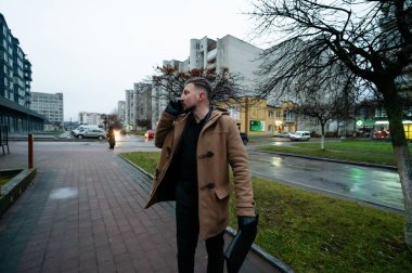 Durgun bir şehir ortamında, zarif ceketli genç bir adam, uzaktaki çağdaş mimari ve ağaçlar tarafından çerçevelenmiş ıslak kaldırım boyunca yürürken telefonuyla konuşur..