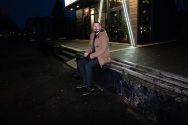 Neon ışıklarının yumuşak parıltıları altında bir adam, gece huzurlu bir şehir ortamında çağdaş bir binanın dışındaki ahşap basamaklara düşünceli bir şekilde oturur..