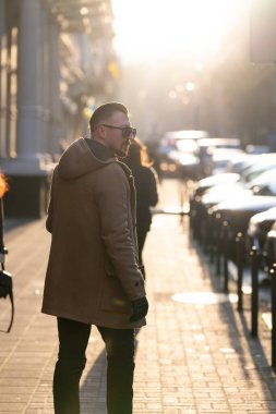 Gün batımının altın ışıltısının ortasında, sıcak palto giymiş şık bir birey canlı bir caddede yürüyor, insanlarla çevrili ve park halindeki arabalarla, mevsimlik cazibesini somutlaştırıyor..