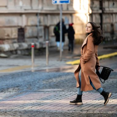 Saçları rüzgarda uçuşan genç bir kadın kaldırım taşları üzerinde zarif bir şekilde yürüyor şık ceketini ve şık şehir ortamında günlük kıyafetlerini sergiliyor..