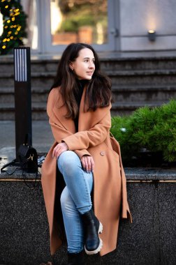 Uzun saçlı genç bir kadın şehir parkında taş basamaklarda sakince oturur. O, yeşil yemyeşil ortamda düşünceli bakarken rahat bir kahverengi ceket ve kot pantolon giyer..